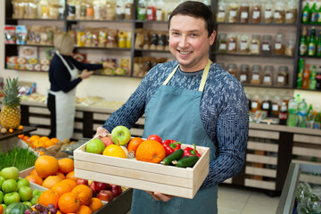 Smiling male salesman holds a wooden box with vegetables and fruits in the store.