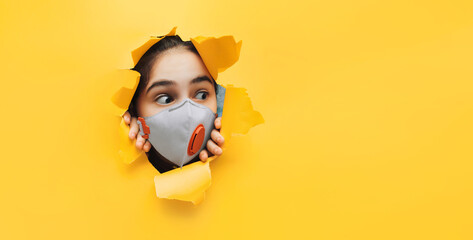 A teenager girl in a white medical mask-respirator looks out curiously from a hole in yellow paper....