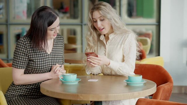 Two charming cheerful young women scanning QR code on restaurant table and talking. Positive Caucasian friends using online menu in cafe indoors. Modern technologies and leisure concept.