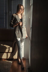 A beautiful blonde in a plaid jacket stands near the window in the rays of the sun passing through the blinds