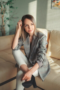 A Beautiful Girl With Blond Hair And Green Eyes In A Plaid Jacket And White Trousers Sits On A Beige Sofa In A Room With Light Green Wallpaper