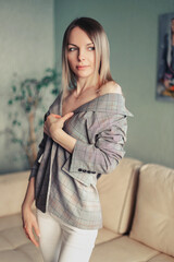 Fototapeta premium A beautiful girl with blond hair and green eyes in a plaid jacket stands and looks out the window in a room with light green wallpaper