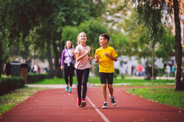 Obraz premium Active recreation and sports children in pre-adolescence. Caucasian twins boy and girl 10 years old jogging on red rubber track through park. Children brother and sister running on treadmill outside