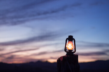An old kerosene lamp lit with the sunset sky and mountains in the background. Sky with clouds and...