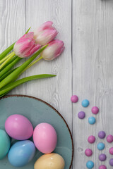 Obraz na płótnie Canvas Pastel colored Easter eggs and pastel colored chocolate candy in a Blue colored plate and pink white tulips, against white wooden background.