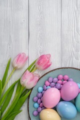 Obraz na płótnie Canvas Pastel colored Easter eggs and pastel colored chocolate candy in a Blue colored plate and pink white tulips, against white wooden background.