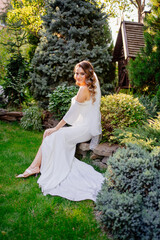 A beautiful bride with long curls sits on a park bench among grass and trees.