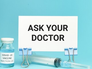 Phrase ASK YOUR DOCTOR written on white card with a bottle of Covid 19 vaccine and syringe.