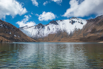 Shore of the lake with rocks and blue water with a background of rocky mountains and partially covered with snow on a sunny morning with blue sky