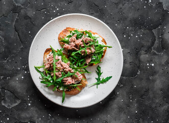 Snack sandwiches with canned tuna, cream cheese and arugula on a dark background, top view