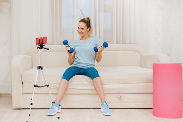 Fototapeta na wymiar Young female fitness trainer with dumbbells recording video blog, work as a trainer from home online remote, recording a video lesson, modern technologies and quarantine