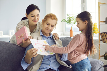 Cheerful grown up daughter holding gift box and granddaughter with drawing greeting senior...