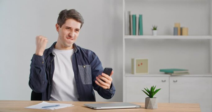 Caucasian male businessperson millennial freelancer sitting at desk finishing up phone call selling online using smartphone making victory gesture receiving good news rejoicing over success triumph