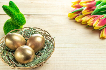 Golden easter colour eggs in basket with spring tulips, white feathers on wooden background in Happy Easter decoration. Spring holiday concept.