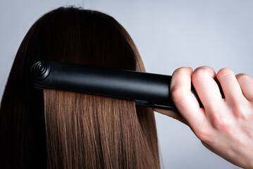 Fototapeta premium Young girl is doing her hair using a straightener on gray background.