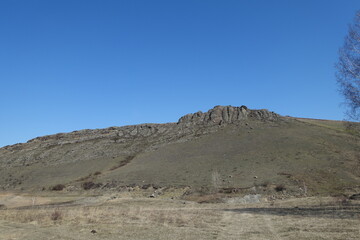 A field and a rocky mountain in early spring against a blue sky. A gray and bleak spring landscape in the countryside.