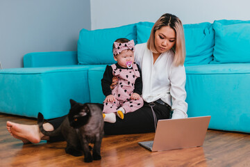 A young mother works over the Internet. A woman of Asian appearance uses a laptop, holding a baby in her arms, sitting on the floor next to a cat.