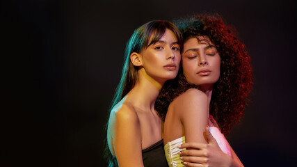 Two stylish young women with professional art makeup posing together in neon light isolated over black background