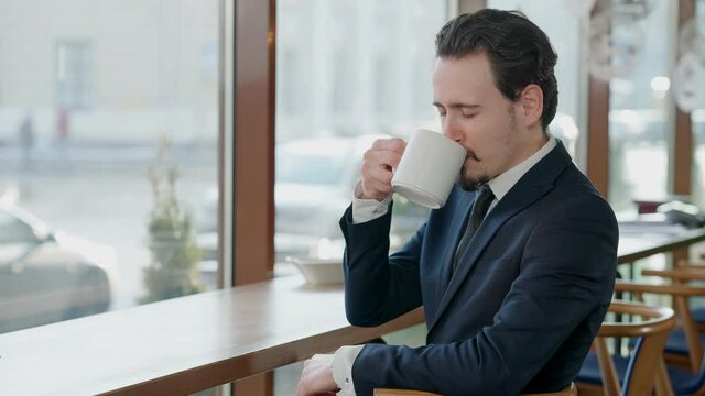 Confident positive businessman posing with tea cup in cafe. Portrait of elegant handsome Caucasian man with beard and mustache drinking healthful hot drink sitting at window on rainy day.