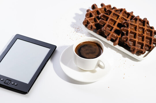 freshly brewed black natural coffee in a cup and with chocolate waffles for breakfast on a white background in a sunny morning, reading on an electronic reader