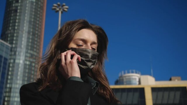 Brunette girl in black antibacterial mask talks on the phone and looks at the clock against the backdrop of the metropolis. High quality FullHD footage