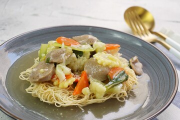 A plate of I Fu Mie or Crispy Fried Noodle with thick savory sauce and meatball and vegetables in shiny and grainy white background. Selective focus