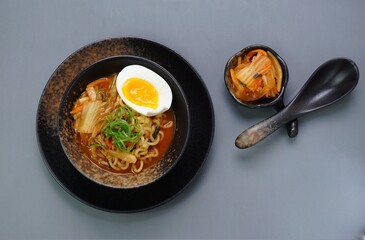 Top view of instan ramyeon or Korean noodles soup on black bowl, topped with half boiled egg, leek and Kimchi on small bowl. Isolated gray background with spoon, napkin and wooden board