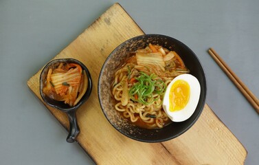 Top view of instan ramyeon or Korean noodles soup on black bowl, topped with half boiled egg, leek and Kimchi on small bowl. Isolated gray background with spoon, napkin and wooden board