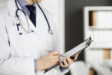 Unknown male doctor standing and working with clipboard of medication history records in clinic at his working place, closeup. Perfect medical service in hospital. Medicine and healthcare concept