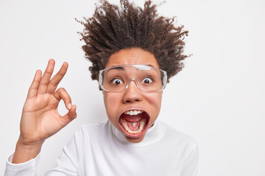Headshot of shocked emotive curly haired young woman stares at camera has jaw dropped raised hair shows okay gesture says I like it has crazy look isolated over white background. Madness concept
