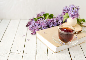 Fototapeta na wymiar Romantic background with a cup of tea, lilac flowers and a book over a white wooden table. Leisure concept, spring breakfast.