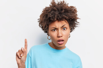 Headshot of displeased curly haired Afro American woman looks puzzled at camera indicates index finger above shows something amazing dressed in casula blue t shirt isolated over white background