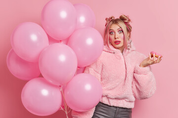 Surprised fashionable woman dressed in stylish clothes makes hairstyle and applies makeup for special occasion holds glazed donut bunch of inflated balloons enjoys holiday poses indoor alone