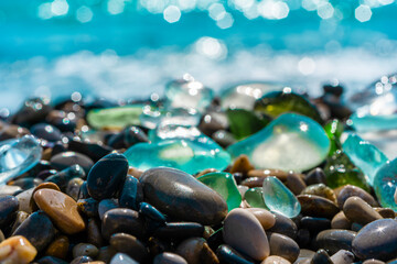 Natural polish textured sea glasses and stones on the seashore. Azur clear sea water with waves. Green, blue shiny glass with multi-colored sea pebbles close-up. Beach summer background