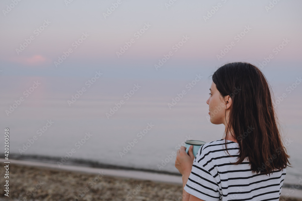 Wall mural young pretty woman drinking hot morning tea looking at sunrise at sea