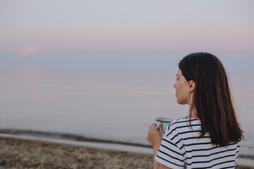 young pretty woman drinking hot morning tea looking at sunrise at sea