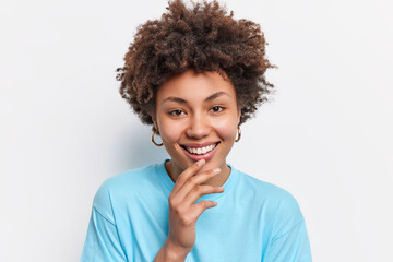 Fototapeta na wymiar Portrait of young beautiful African American keeps hand on chin smiles gently looks directly at camera dressed in blue t shirt expresses positive emotions isolated over white studio background