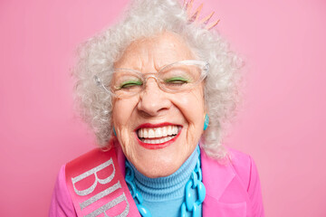 Close up shot of positive wrinkled old woman smiles toothily at camera wears transparent glasses...