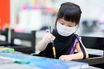 Asian girl wearing white mask prevents the spread of virus. Children learn art in class. Child makes crafts on canvas and wear black apron to prevent water from getting stained with his clothes.
