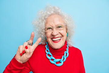 Portrait of cheerful nice looking elderly woman smiles happily makes peace gesture shows v sign...