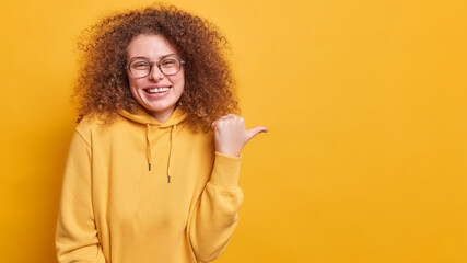 Glad positive curly haired woman in eyeglasses smiles sincerely advertises product has good mood demonstrates copy space points with thumb dressed in sweatshirt isolated over yellow background