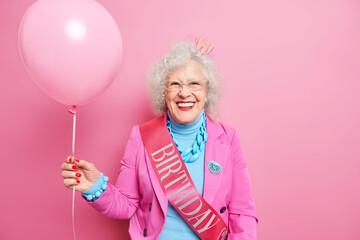 Isolated shot of beautiful wrinkled woman smiles positively celebrates anniversary has fun on party for elderly people wears festive clothes with ribbon holds inflated balloon enjoys birthday