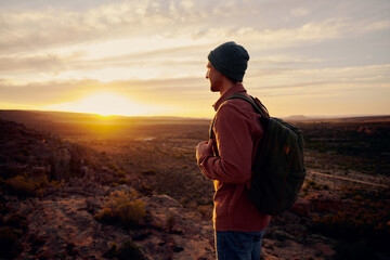 Happy tourist man with backpack standing on top mountain looking into the distance at morning sunrise