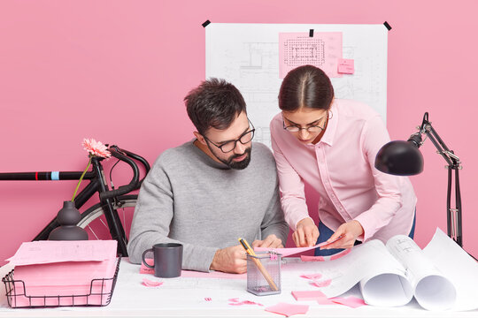 Indoor shot of busy woman and man work together on design project pose in coworking space. Two architects discuss new house plans sit at office desk with blueprints around. Collaboration concept