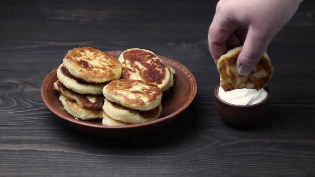 Cheesecakes on a clay plate and a bowl of sour cream. A hand dips a cheesecake in the cream