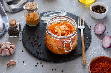 Spicy Korean appetizer kimchi made of cabbage with carrots in a glass jar with spices garlic and onions on a dark dish. Horizontal orientation, gray background, no people.