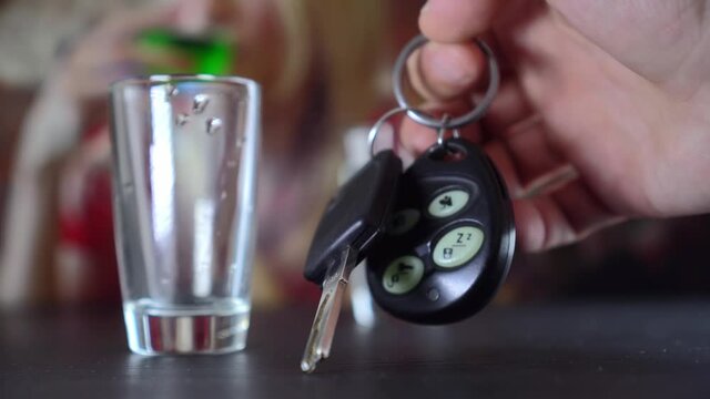 On black table are car keys and glass of vodka. man's hand takes glass, drinks, takes car keys. A noisy party, bar, club or disco. In background, woman drinks absinthe. Driving without alcohol.