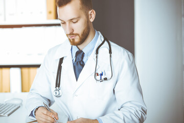 Friendly red-bearded doctor sitting and writing at clipboard in sunny clinic. Medicine concept, pandemic stop