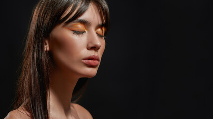 Fototapeta premium Close up beauty portrait of young brunette woman with colorful orange makeup posing with eyes closed isolated over black background