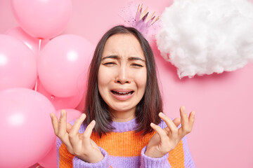 Upset lonely woman cries from despair sad to celebrate birthday alone raises hands from frustration wears princess crown and jumper prepared for festive party poses against decorated background